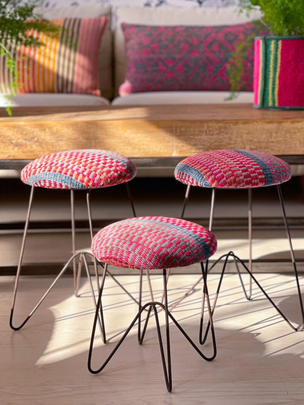 Español: Tres reposapiés con tapizado en tejido étnico que presenta un patrón geométrico en colores rosados y azules, con bases de metal negro de cuatro patas en forma de horquilla. English: Three footrests upholstered in ethnic fabric that presents a geometric pattern in pink and blue colors, with black metal bases with four hairpin-shaped legs.