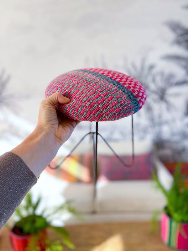 ESPAÑOL: Parte superior de un reposapiés étnico de tela con un patrón de tejido en rosa y azul. El reposapiés tiene una base de alambre negra de tres patas. INGLES: Top of an ethnic fabric footstool with a woven pattern in pink and blue. The footstool has a three-legged black wire base.