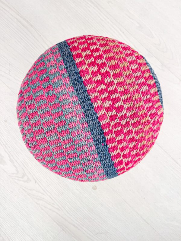 ESPAÑOL: Vista aérea de un reposapiés étnico tejido, con una mitad en tonos rosas y la otra en azules y grises, sobre un suelo de madera blanca. ENGLISH: Aerial view of a woven ethnic footstool, with one half in pink tones and the other in blues and grays, on a white wooden floor.